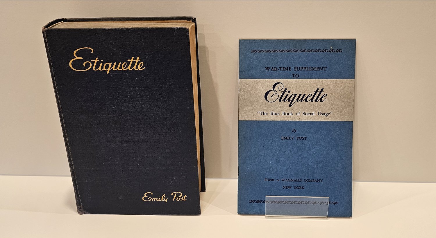 A dark hardcover copy of Emily Post's Etiquette beside a slim blue War-Time Supplement booklet.